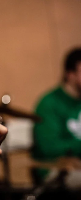 A close-up of a Fender guitar headstock, with a drummer in a green hoodie slightly out of focus behind it. A close-up of a Fender guitar headstock, with a drummer in a green hoodie slightly out of focus behind it.
